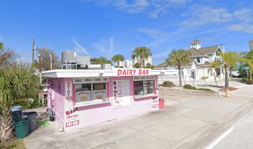 A pink dairy bar with a menu, surrounded by palm trees and a clear blue sky.