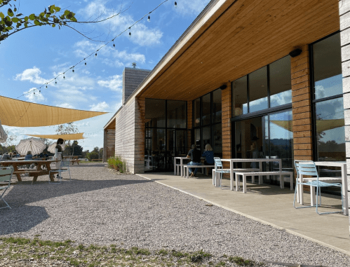A modern building with large windows, outdoor seating, and string lights, set in a sunny landscape.