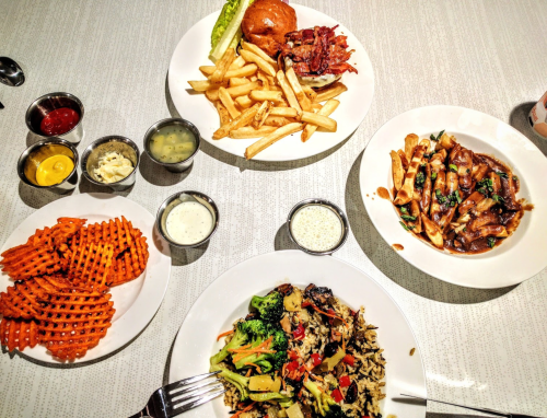 A table with various dishes: a burger with fries, pasta, stir-fried vegetables, and waffle fries with dipping sauces.