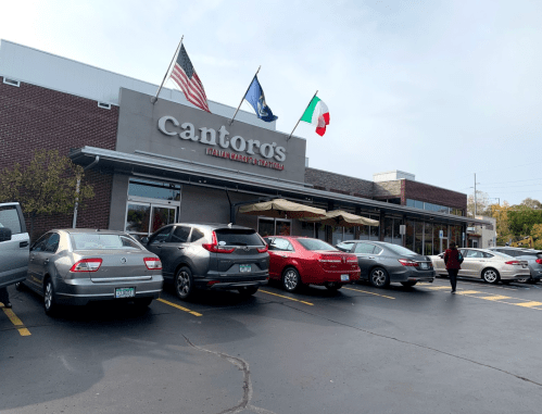 Exterior of Cantoro's Italian Market with flags and parked cars in the lot on a clear day.