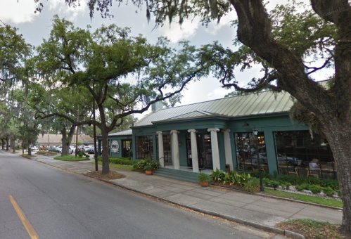 A green building with columns and large windows, surrounded by trees, along a quiet street.