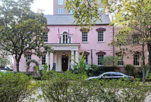 A pink historic house with flags, surrounded by greenery and parked cars, showcasing classic architectural features.