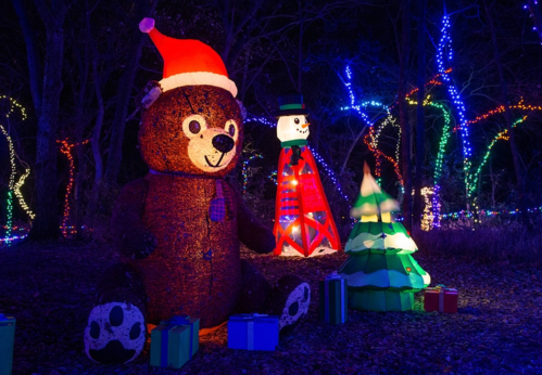 A large teddy bear in a Santa hat sits among colorful holiday lights, with a snowman, Christmas tree, and presents nearby.