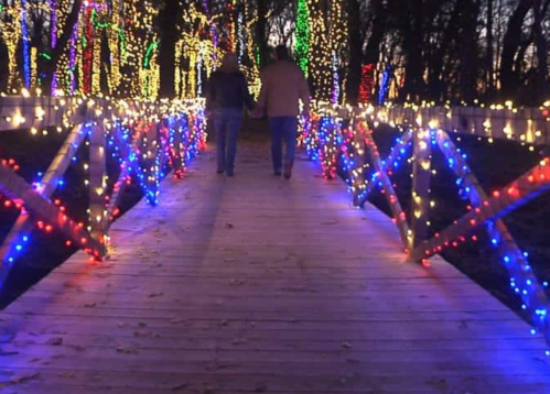 A couple walks hand in hand on a lit bridge surrounded by colorful holiday lights in a wooded area.