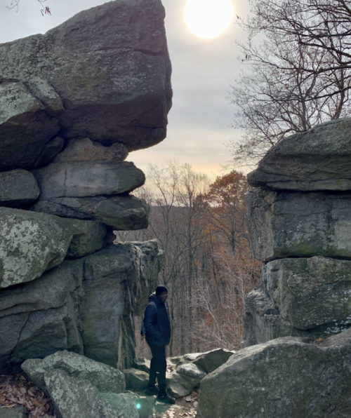 A person stands between large rock formations, with a sunlit sky and trees in the background.