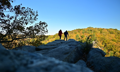 Two hikers silhouetted against a clear blue sky, walking on a rocky outcrop with autumn foliage in the background.