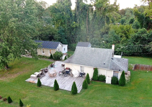 Aerial view of a house with a wooden deck, surrounded by trees and a grassy yard, with another building nearby.