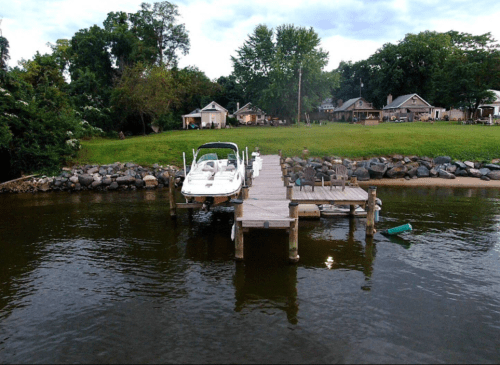 A boat docked at a wooden pier by a calm river, with houses and trees in the background.