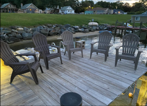 A wooden dock by the water with five gray chairs arranged in a circle, surrounded by houses and greenery.
