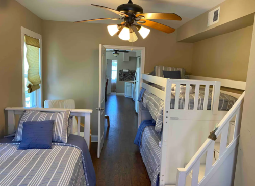 A cozy room with a ceiling fan, featuring a bunk bed and a double bed, with light-colored walls and wooden flooring.