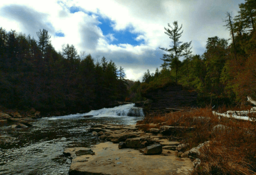 A serene river flows through a forested landscape, with rocky banks and a waterfall under a cloudy sky.
