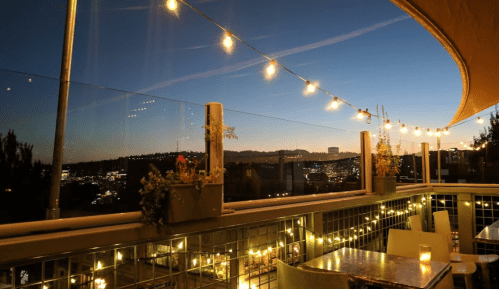 A rooftop terrace at dusk, adorned with string lights and potted plants, overlooking a city skyline.