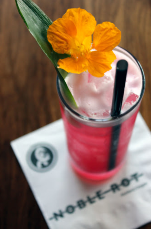 A vibrant pink cocktail garnished with a yellow flower and a green leaf, served on a napkin with "Noble Rot" printed on it.