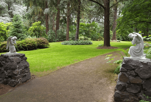 A serene garden path flanked by stone sculptures of angels, surrounded by lush greenery and trees.