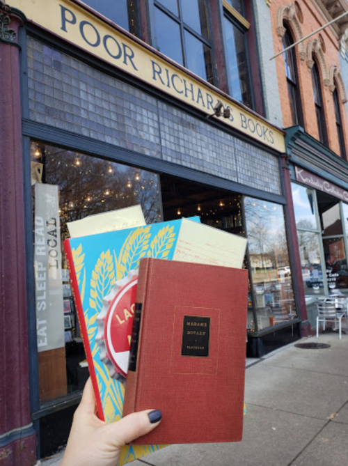 A hand holds three books in front of Poor Richard's Books, a charming bookstore with a vintage facade.