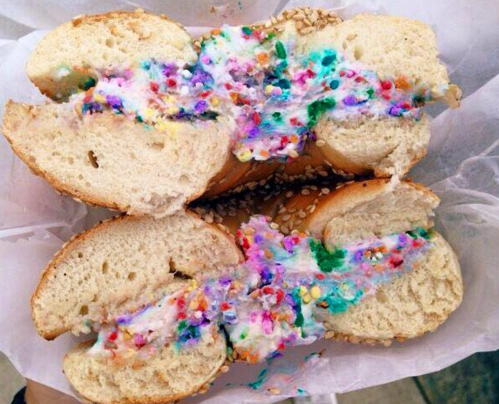A split bagel filled with colorful cream cheese and sprinkles, resting on white paper.