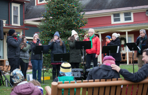 A group of singers performs holiday music outdoors near a decorated tree, with an audience seated on benches.