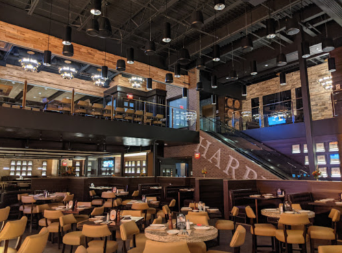 Interior of a modern restaurant featuring wooden accents, round tables, and a staircase leading to an upper level.