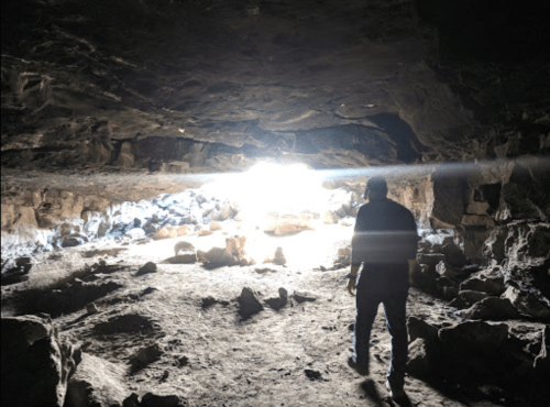 A person stands in a dark cave, facing a bright light at the entrance, surrounded by rocks and shadows.