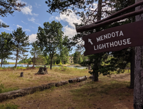 A wooden sign points to Mendota Lighthouse, surrounded by trees and a serene lake under a partly cloudy sky.