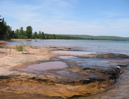 A serene lakeside view with sandy shores, rocky outcrops, and lush green trees under a clear blue sky.
