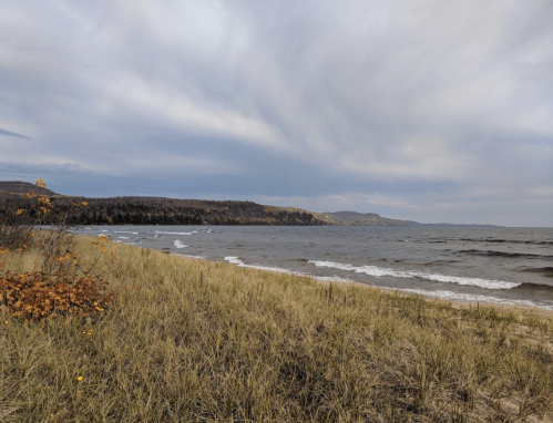 A serene beach scene with gentle waves, grassy shore, and distant hills under a cloudy sky.