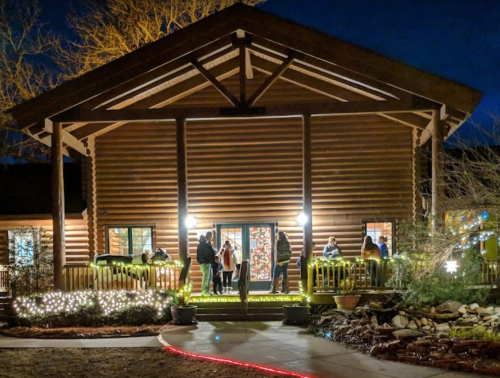 A log cabin at night, illuminated with lights, with people gathered on the porch and around the entrance.