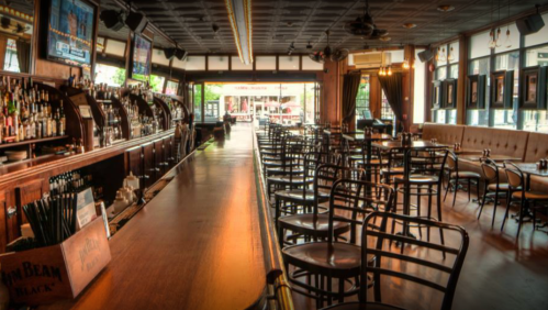 A spacious bar interior with a long wooden counter, empty tables, and large windows letting in natural light.