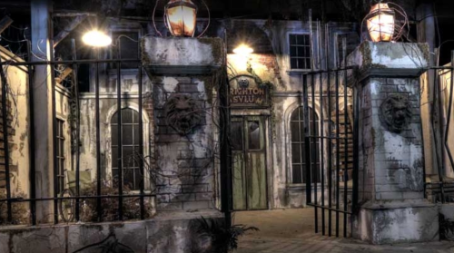 A dark, eerie entrance to an abandoned asylum, featuring stone pillars, barred gates, and dim lanterns.