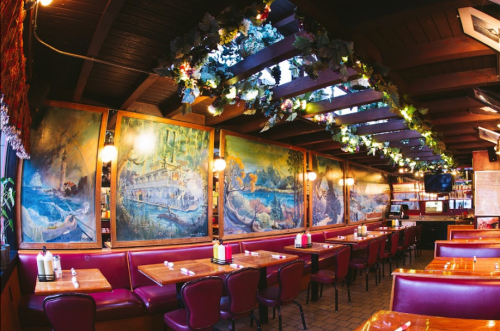 Cozy restaurant interior with wooden beams, vibrant murals, and festive greenery above red booths and tables.