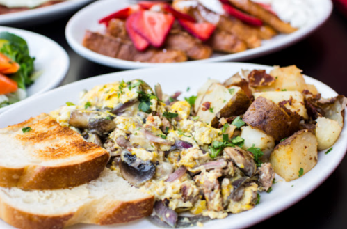 A plate of scrambled eggs with mushrooms, potatoes, and toast, with a side of salad and other dishes in the background.