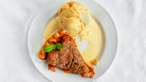 A slice of pie with a scoop of ice cream, drizzled with sauce, garnished with a mint leaf on a white plate.
