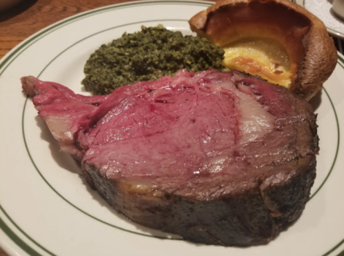 A plate featuring a large slice of rare prime rib, a serving of green spinach, and a Yorkshire pudding.