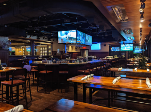 Interior of a modern restaurant with wooden tables, bar seating, and multiple screens displaying sports.
