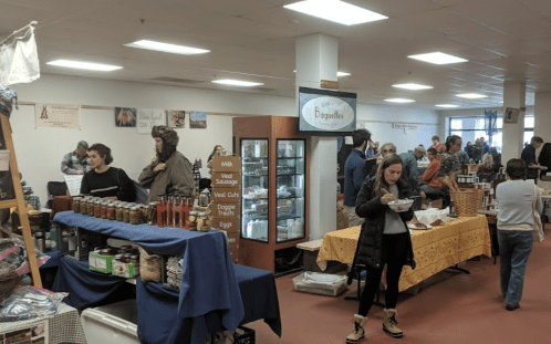 A bustling market scene with people browsing stalls, shopping for local goods, and enjoying food in a bright indoor space.
