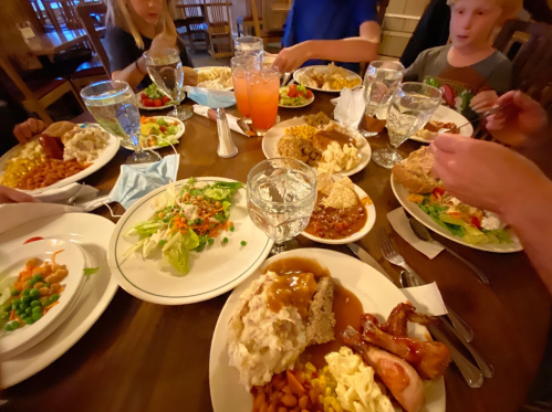 A table filled with various dishes, including salads, meats, and drinks, surrounded by people enjoying a meal together.