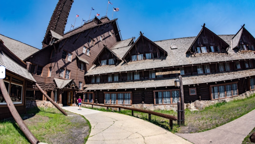 A large, rustic wooden lodge with multiple stories, surrounded by green grass and a clear blue sky.