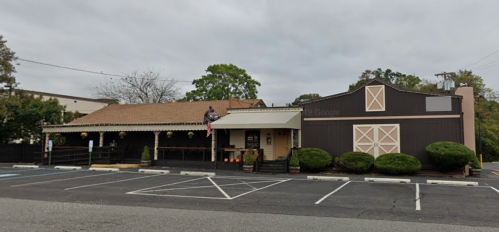 A rustic restaurant with a brown exterior, porch, and decorative plants, set in a parking lot on a cloudy day.