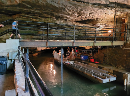 A dimly lit cave interior with a walkway, water, and people near a boat docked at the edge.
