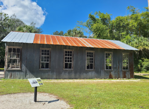 A weathered metal building with a rusty roof, surrounded by greenery and a sign in front on a sunny day.