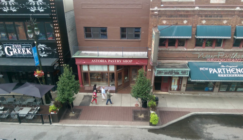 A street view of Astoria Pastry Shop with pedestrians walking by and nearby restaurants in a bustling urban area.