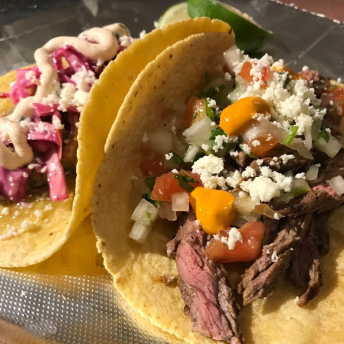 Two tacos on a silver tray: one with beef, diced tomatoes, and cheese; the other with pickled onions and sauce.