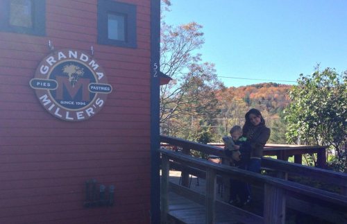 A woman holds a child on a wooden walkway beside a red building with a sign for "Grandma Miller's Pies and Pastries."