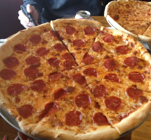 A large pepperoni pizza with several slices cut, sitting on a wooden table next to another pizza and a soda can.