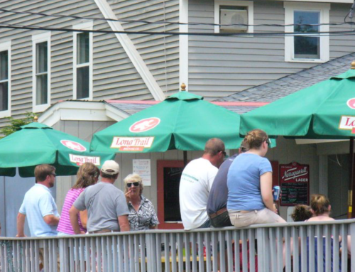 A group of people socializing under green umbrellas at an outdoor venue, with a building in the background.