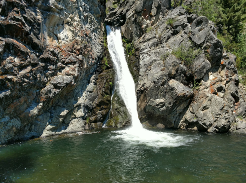 A waterfall cascades down rocky cliffs into a clear pool surrounded by lush greenery.