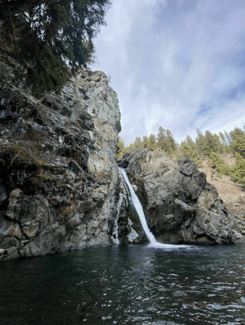 A serene waterfall cascading down rocky cliffs into a clear pool, surrounded by lush green trees and a cloudy sky.