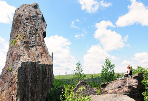 A person sits on a rock, gazing up at a large boulder under a blue sky with scattered clouds and greenery around.