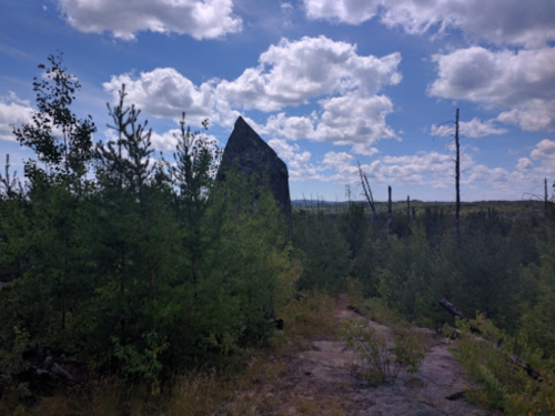 A tall rock formation stands amidst green trees and a blue sky with scattered clouds.