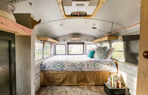 Cozy interior of a converted bus, featuring a bed with decorative pillows, wooden accents, and natural light.
